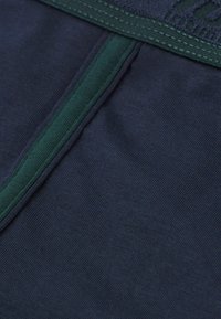 Mörkblått tyg med en slät textur, med en grön accentremsa och en sydd midjeband. Närbild som betonar sömmarnas kvalitet.