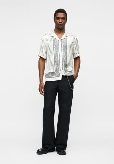 Άνδρας που στέκεται, φοράει ένα λευκό κοντομάνικο πουκάμισο με σχέδιο, μαύρο παντελόνι με αλυσίδα και μαύρα παπούτσια πάνω σε απλό λευκό φόντο.
