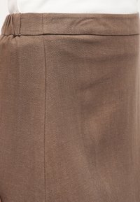 Κοντινό πλάνο προσώπου που φορά καφέ παντελόνι από λινό με ελαστική μέση και λευκό πουκάμισο, που έχει μπει μέσα στο παντελόνι.