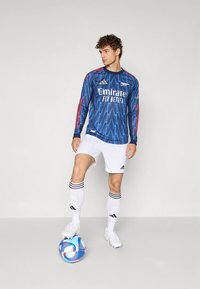 Niebieska piłkarska koszulka z długim rękawem z wzorem w zygzak, czerwone akcenty, białe spodenki, czarno-białe paskowane skarpetki oraz kolorowa piłka nożna.