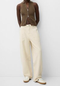 Personne portant un haut sans manches en maille marron avec fermeture éclair, un pantalon ample couleur crème avec de grandes poches, et des baskets blanches avec des semelles en gomme.