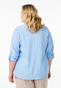 Camicia azzurro chiaro, con bottoni, dal tessuto liscio, maniche arrotolate e orlo arrotondato, abbinata a pantaloni beige.