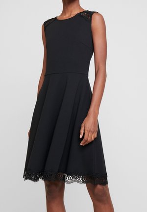 Robe noire sans manches avec détails en dentelle aux épaules et à l'ourlet, corsage ajusté et jupe évasée portée par une personne debout.