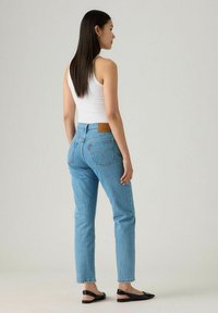 Jeans de mezclilla azul claro con corte recto, que presentan bolsillos traseros y un parche de cuero marrón. Combinados con una camiseta sin mangas blanca y bailarinas negras.