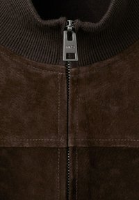 Gros plan d'une veste en suède marron avec col côtelé et tirette de fermeture éclair métallique gravée "MNG" au centre.