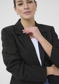 Schwarze Blazer mit Reverskragen, strukturiertem Stoff und zwei Seitentaschen. Getragen über einem weißen Oberteil mit strukturierter Passform und umgeschlagenen Ärmeln.
