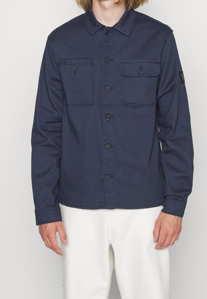 Homme portant une chemise bleu marine boutonnée avec deux poches poitrine et un pantalon blanc, debout devant un fond uni.
