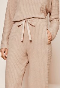 Conjunto de loungewear de canales beige que presenta una cintura con cordón, mangas largas y bolsillos laterales. Tejido suave con una textura de brillo sutil.