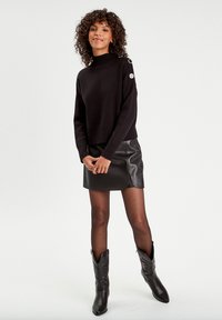 Pull en tricot noir avec col montant et accents de boutons argentés, associé à une mini-jupe en simili cuir noir et des bottes de cow-boy noires jusqu'aux genoux.