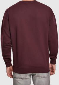Uomo che indossa un semplice sweatshirt a maniche lunghe color bordeaux e jeans grigi chiari, visto di spalle su uno sfondo bianco.