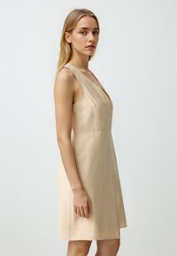 Korte beigekleurige jurk met een v-hals en sans-mouwen ontwerp. Gemaakt van gladde stof, heeft het een getailleerde taille en een uitlopende rok.
