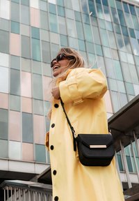 Geel waterdichte regenjas met zwarte knopen en een zwarte schoudertas met een textuur in goudaccent. Gebouw met glazen panelen op de achtergrond.
