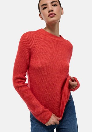 Pull en maille rouge avec un col rond, texture côtelée et manches longues. Une coupe décontractée et un ourlet replié ajoutent du détail. Associé à un jean bleu.