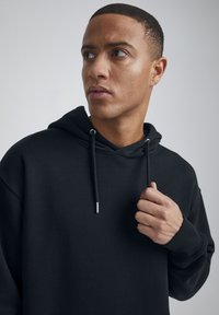 Sudadera negra hecha de un tejido suave, con capucha ajustable, puños de canalé y un corte relajado. El diseño es sencillo y minimalista.