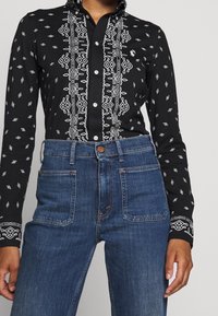 Svart skjorta med knappar och vit geometrisk broderi längs framsidan och ärmarna, tillsammans med högmidjade blå denimjeans.