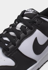 Κοντινό πλάνο μαύρου και άσπρου αθλητικού παπουτσιού Nike, εστιασμένο στην μπροστινή δέσιμο κορδονιών, τις ραμμένες λεπτομέρειες από δέρμα και το εμφανές λογότυπο Nike στην πλευρά.