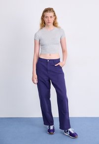 Grijze cropped t-shirt, paarse losse broek met voorzakken, en paarse sneakers met lichtblauwe accenten op een blauwe achtergrond.