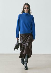 Frau, die einen blauen Rollkragenpullover, einen braunen Lederwickelrock, schwarze Stiefel, Sonnenbrille trägt und eine kleine schwarze Handtasche hält.
