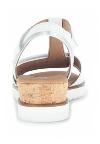 Sandale blanche avec des accents de sangle métallique et un talon en liège. Semelle flexible, dos ouvert et bride de cheville ajustable pour un maintien sécurisé.