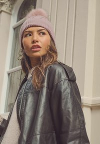 Kootud roosa beanie karvase pom-pomiga, koos musta nahktagi ja heledate seintega taustaga, kus on aknad.