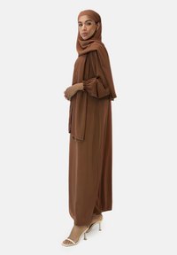 Brun abaya lavet af glat stof; har flydende ærmer og sideslids. Bæres med matchende hijab og stropheels. Elegant silhuet.