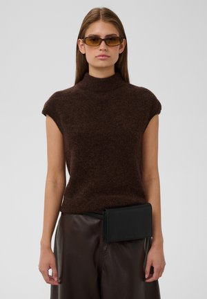 Mujer con jersey marrón sin mangas, pantalones marrón oscuro, gafas de sol y un bolso negro de cinturón sobre un fondo gris liso.