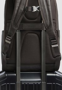 Bruine rugzak met structuur, verstelbare bandjes en metalen beslag, geplaatst op een zwarte koffer met wieltjes en een geribbelde oppervlakte.