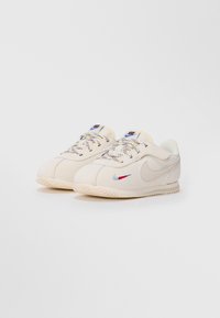 Par zet beige Nike tenisica s višebojnim vezicama, malim crvenim i plavim izvezenim swoosh-om, i bež teksturiranim đonom na bijeloj pozadini.