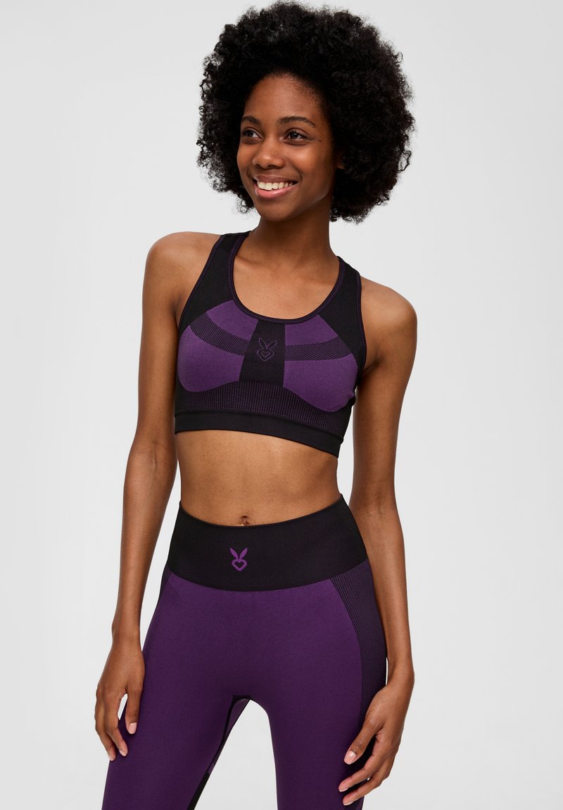 Frau, die lächelt, trägt einen nahtlosen Sport-BH in Lila und Schwarz sowie passende hochgeschnittene Leggings, steht vor einem schlichten hellen Hintergrund.