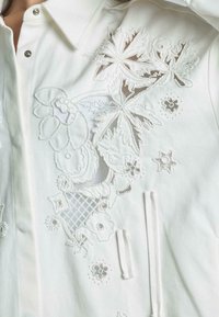 Chaqueta de mezclilla blanca con intrincados bordados florales en blanco y acentos de cuentas, acabado texturizado, cierre a presión y cordón ajustable.
