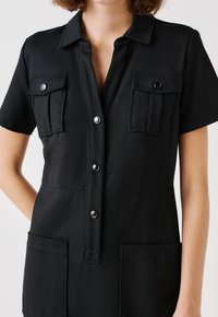 Schwarzer Kurzarm-Overall aus glattem Stoff mit einem kragenlosen Ausschnitt, vorderer Knopfleiste und zwei Brusttaschen mit Druckknöpfen.