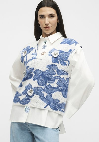Femme portant une chemise blanche à col et un gilet en maille blanc avec un grand motif floral bleu et des fleurs décoratives, associée à un jean bleu clair.