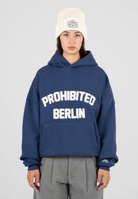 Sudadera con capucha azul marino con "PROHIBIDO BERLÍN" en letras blancas, con un bolsillo delantero y puños acanalados. Combinada con pantalones grises con pliegues.