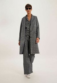 Ensemble en deux pièces avec un motif géométrique bleu marine et blanc ; manteau long et pantalon ; le tissu semble lisse ; associé à des chaussures blanches et des lunettes de soleil.