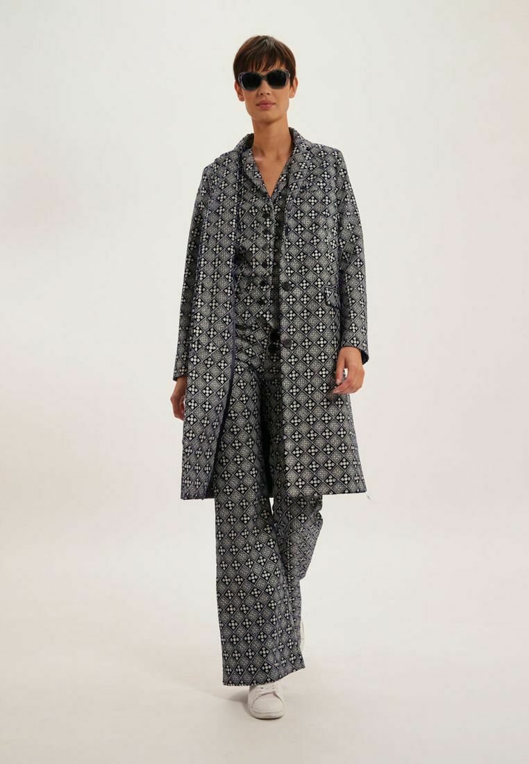 Ensemble en deux pièces avec un motif géométrique bleu marine et blanc ; manteau long et pantalon ; le tissu semble lisse ; associé à des chaussures blanches et des lunettes de soleil.