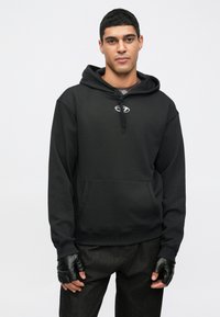 Sweatshirt preta com capuz e bolso frontal, apresenta um pequeno logótipo no peito, combinada com jeans escuros e luvas sem dedos.