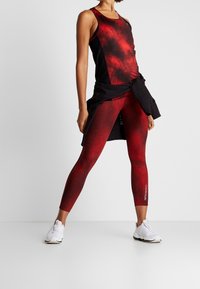 Rød og sort atletisk tanktop med et spraglet design, parret med matchende højlivede leggings. Hvide atletiske sko.