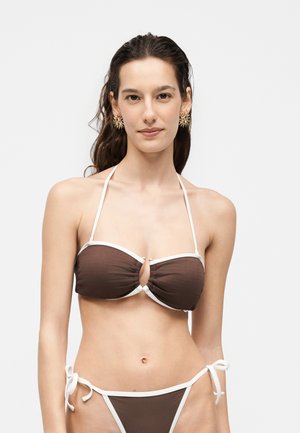 Giovane donna con lunghi capelli scuri che indossa un bikini marrone con bordo bianco e orecchini a forma di sole dorati, in piedi su uno sfondo chiaro.