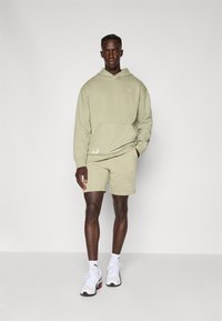 Hoodie en coton vert olive avec poche kangourou, associé à un short assorti. Présente un logo en crème, complété par des baskets blanches.