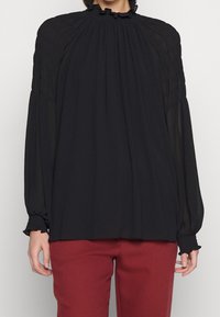 Blusa nera a maniche lunghe con un colletto alto e arricciato, caratterizzata da una texture arricciata lungo le spalle e i polsini, abbinata a pantaloni bordeaux.