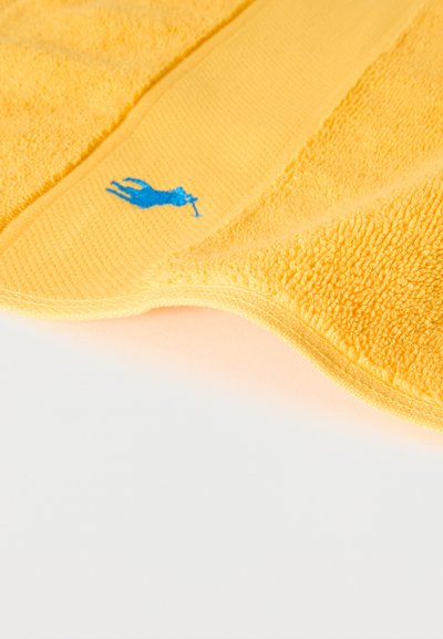 Κίτρινη πετσέτα από περκάλι με κέντημα λογότυπου παίκτη πόλο σε μπλε χρώμα κατά μήκος μιας άκρης σε λευκό φόντο.