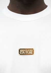 Άτομο με σκούρο χρώμα δέρματος που φοράει λευκό πουκάμισο με ένα χρυσό ορθογώνιο σήμα στο στήθος που γράφει "VERSACE JEANS COUTURE".