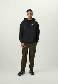 Zwarte hoodie met een voorzak, gecombineerd met donkerolijfgroene sweatpants met rode en zwarte accenten aan de zijkanten. Grijze sneakers.