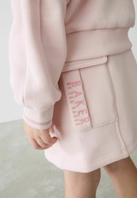 Gaiši rozā sporta krekls un svārku komplekts ar ribotām piedurknēm, kuram ir kabata ar izšuvumu "BAKER by Ted Baker" atbilstošā rozā krāsā.