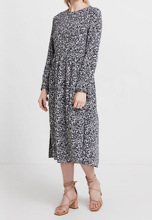 Woman wearing a long-sleeve, black and white floral midi dress with beige strappy block-heeled sandals standing on a plain background.