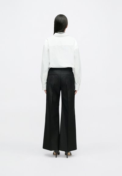 Witte blouse met lange mouwen en een achterpas, gecombineerd met een zwarte, hooggeposeerde broek met wijde pijpen en hakken, gezien van achteren.