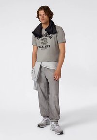 T-shirt gris en coton avec logo "Champion" noir, associé à un pantalon texturé gris clair et des baskets grises. Une serviette noire est posée sur les épaules.