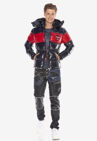 Puffjacke mit glänzenden navyblauen und roten Abschnitten, Kapuze, Reißverschlusstaschen. Kombiniert mit dunklen Jeans, die verschiedene Texturen, Muster und Nähte aufweisen.