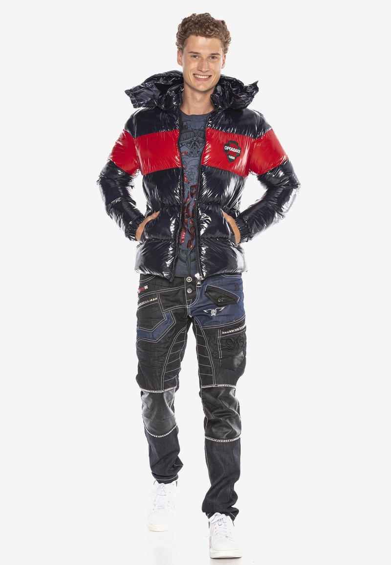 Puffjacke mit glänzenden navyblauen und roten Abschnitten, Kapuze, Reißverschlusstaschen. Kombiniert mit dunklen Jeans, die verschiedene Texturen, Muster und Nähte aufweisen.