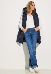 Gilet rembourré marine avec capuche, associé à une chemise blanche à manches longues et un jean bleu, complété par des baskets blanches. Design simple et coupe décontractée.